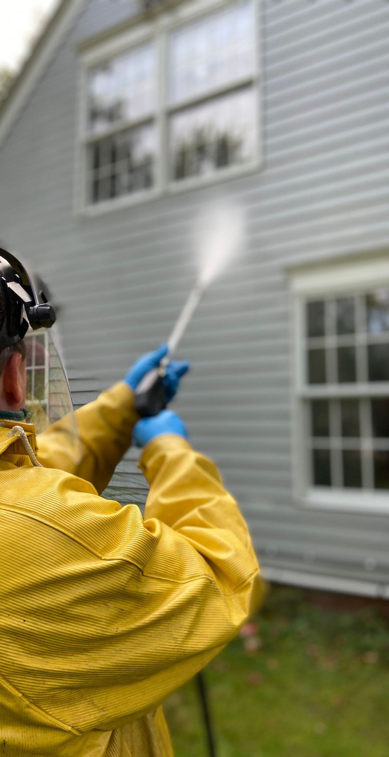Person cleaning house siding with a pressure washer, wearing protective gear in a yellow raincoat.